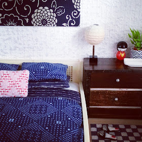 Corner of a one-twelfth scale Japanese-themed bedroom with a cream cane bed with blue-and-white bedding, a chest of drawers with a rice-paper lamp. a kokeshi dolls, a plant in a blue-and-white Japanese pot and a white rectangular bowl displayed on it and, on the floor, a blue and white checked rug with a pair of embroidered slippers on it.