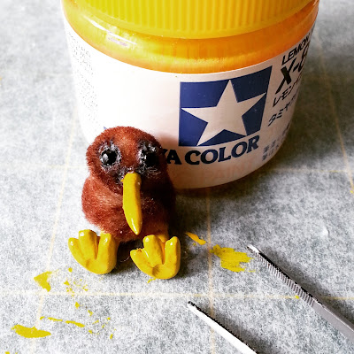 One-twelfth scale kiwi soft toy sitting against a full-sized jar of yellow paint, next to a pair of tweezers.