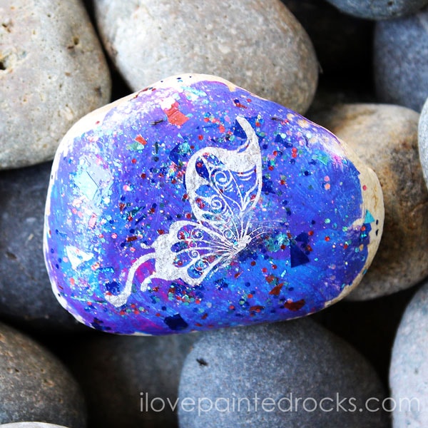 Rock painting tutorial: How to make a glitter butterfly painted rock. This is such a pretty rock painting idea! I l love the galaxy effect of the glitter together with the metallic butterfly from the temporary tattoo. #ilovepaintedrocks #rockpainting #paintedrocks #easycraft #kidscraft #rockpaintingideas