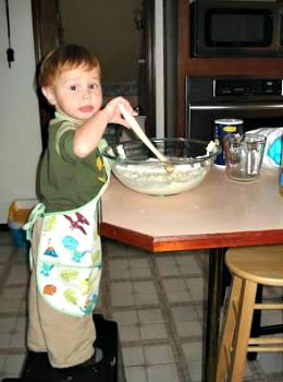 A young boy wearing an apron and standing at a table, stirring batter with a spoon.