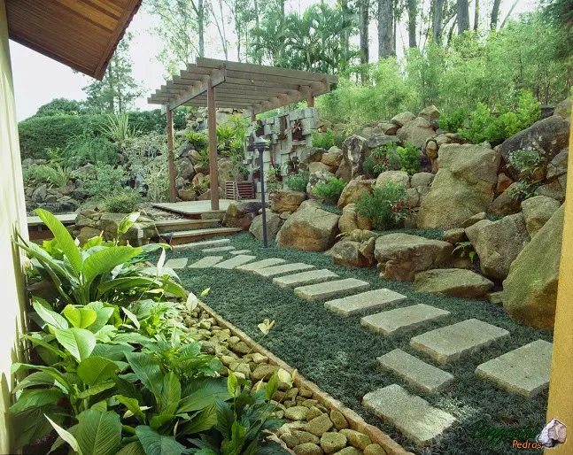 Caminho com pedra no jardim, muro de pedra artesanal com o pergolado de madeira e a canaleta com pedra moledo para captar a água da chuva.
