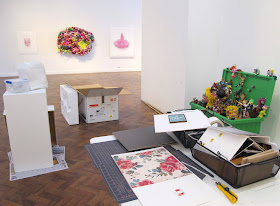 View across a gallery during install, showing a table with a miniature scene being put together and various artworks in the background, mounted or still in packaging.