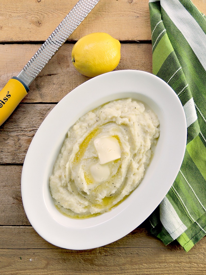 Lemon Garlic Mashed Cauliflower "Potatoes" topped with a pat of butter in a white bowl on a wooden background. Lemon and zester with a green towel on the side.