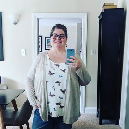 image of me from mid-thigh up, standing in a mirror taking a picture of myself, with my hair up, wearing glasses, a beige cardigan, a white blouse with a colorful butterfly pattern on it, and blue jeans