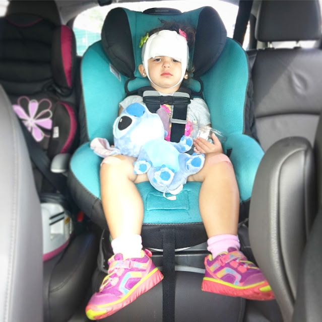 Sad toddler sits in her carseat with stuffed animal in her hand and bandages tied around her head