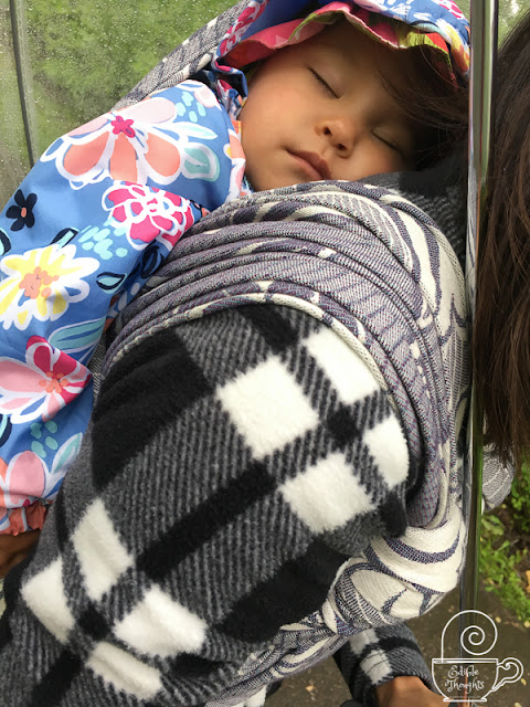 [Image of a peacefully sleeping toddler wrapped on Mama's back in a grayish purplish geofloral woven wrap. Toddler has on a blue floral raincoat. Mama has on a black and white plaid fleece. Mama is holding on to a clear domed umbrella. A wrap nap out on a walk like this is heavenly.]
