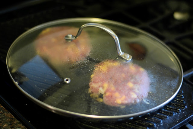A pan lid over the bacon cheeseburger patties on the grill pan.  