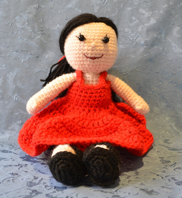 Kwokkie Doll seated in her red sundress, front view. Her straight black hair is tied back in a low ponytail with a red ribbon of yarn.