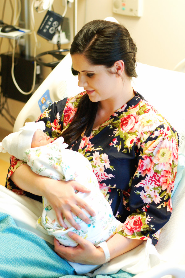 Brunette woman in floral robe holds baby girl in hospital bed after labor.