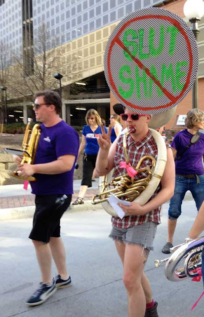 Tuba player with opening of horn covered with handmade sign that says No Slut Shame