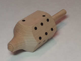 A wooden object shaped somewhat like a dreidel, but with six sides instead of four, and a series of black dots painted on each side in the configuration normally found on regular cubical dice.