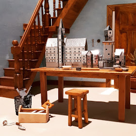 One-twelfth scale miniature room with a worktable in the foreground containing miniature metal model buildings, and a selection of tools.