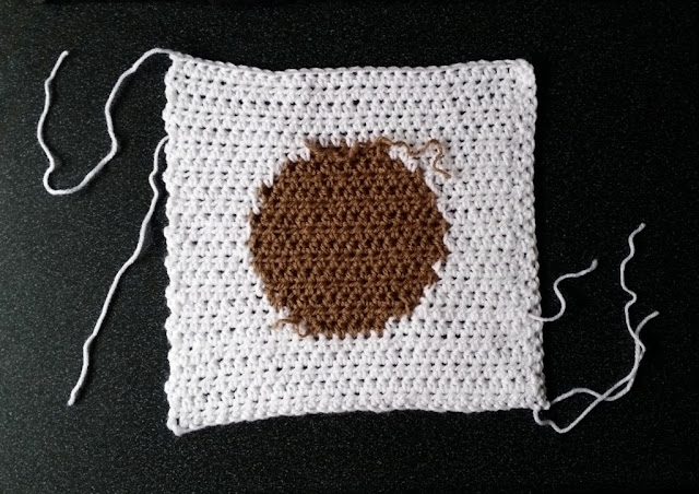 This is the same crocheted square as the previous photograph except it has been flipped over from left to right to reveal the wrong side (or underside) ot the square. Again, the white loose ends can be seen around the edges but now a tan loose end can be seen near the top of the tan circle.  The margins of the circle where the tan and white meet are not as neat as on the right side (or front side) of the square.