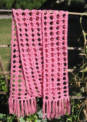 A photograph of a pink lacy scarf hanging over a bamboo trellis with a garden backdrop. The scarf is made using a tricot technique and has straight fringing on each end. 