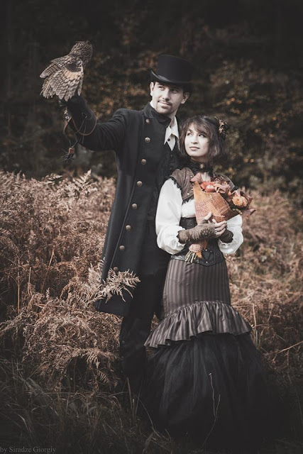 Well dressed Steampunk couple. The woman wears a striped trumpet skirt, corset and bolero jacket. The man wears a long jacket, top hat, and holds a falcon.