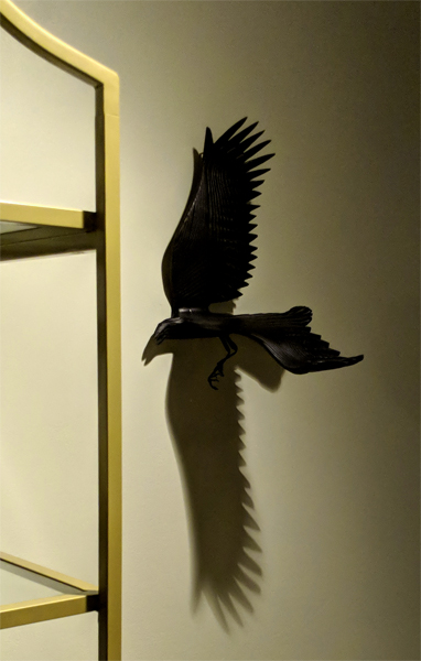 image of a wall-mounted sculpture of a crow, caught in the light in such a way that the wing extended away from the wall is casting a shadow that looks like another wing stretching down the wall
