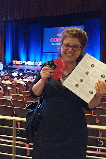 Anna standing in front of TEDx stage "Uncommon", holding speaker and VIP name tags and program with her name