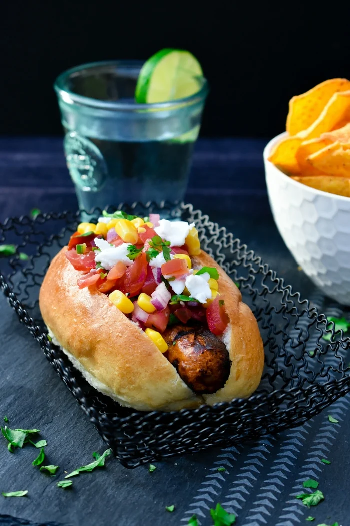 The Mexican Hot Dog - vegetarian hot dog served topped with fresh salsa, corn & coriander, beside a bowl of nachos and a glass of water with a slice of fresh lime in the rim