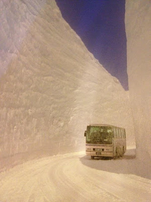 Coach bus driving in snow-packed road between flat-sided snow walls two stories high