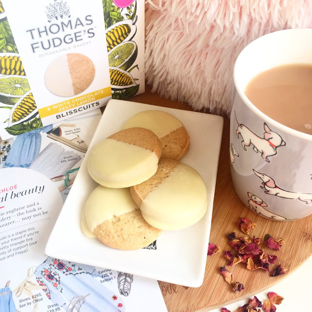 trinket dish with biscuits in the centre on heart wooden chopping board, cup of tea to the right, magazine underneath to the left and box of biscuits in the background