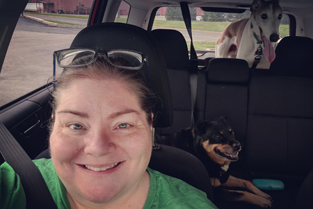 image of me in the car, from the shoulders up, wearing a green t-shirt, with my hair pulled back and glasses sitting on top of my head, smiling; Zelda the Black and Tan Mutt is pictured in the backseat, and Dudley the Greyhound can be seen in the far back compartment of our Subaru Forester