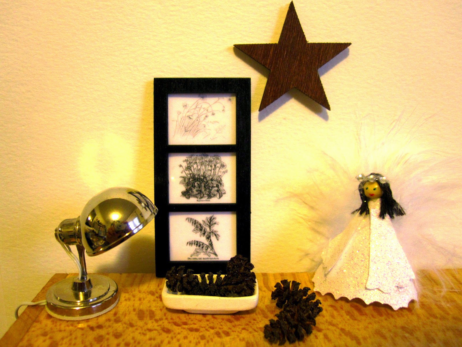 Modern miniature cupboard-top scene which includes a silver lamp, a bowl of pinecones, a three-section picture, a Christmas angel and a wooden star on the wall.