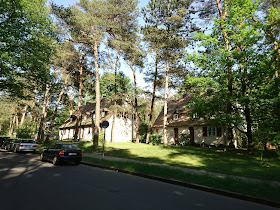 Blick entlang der Straße in der Wohnsiedlung Krumme Lanke. Ein kitschiges Holzartiges Haus steht unter Nadelbäumen.