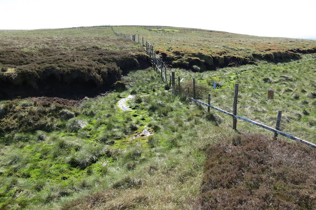 A fence stretches uphill across the moor but the line of the path is broken by a wide bog.