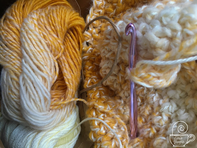 Image of a yellow and cream skin of yarn next to a shimmery cream skein of yarn in a handled brown paper bag nestled close to a crocheted work in progress attached to a pink metal crochet hook.