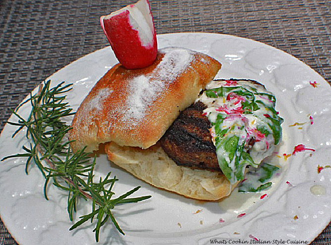 This is a hamburger on a white plate with spinach lemon cream sauce over the top with a sprig of rosemary on the plate on a ciabatta roll with a flowered radish on top