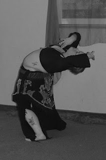 Author in 2009 bellydancing. Photo is black & white, with short hair, black long sleeved crop top, tiger print hip shawl, and black flared pants with tattoo showing on pale skin, performing a standing layback.