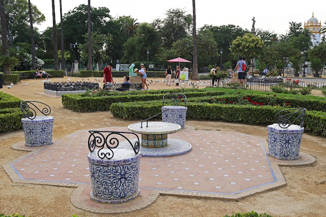 Bancos de azulejos y herrajes entorno a un reloj de sol en un parque con gente y palomas al fondo.