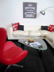 Modern one-tweltfh scale miniature hotel lobby in red, white and black. On the coffee table are a selection of postcards, a glass of wine and a travel magazine.