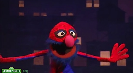 Grover, the fuzzy blue Muppet with big pink nose wearing a Spider-Man costume, including half-face mask, posing with arms out and mouth open against painted city backdrop