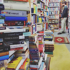 Interior of a second-hand bookshop, with full shelves and stacks of books on the counter.