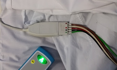 resting on the white sheets of the hospital bed is a white plug with five wires attached to it in brown, white, red black and green.  Below it is the nurse call bell, a blue and grey rectangle with a green button that lights up when callilng for assistance. The small round yellow button will cancel the call.