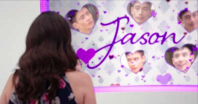 A Janet dressed as Tahani stands with her back towards the camera. In front of her, there's a screen that has "Jason" written on it in loopy letters, and there are a bunch of heart-shaped pictures of Jason's face.