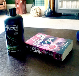 A black large cylindrical bottle with a cylindrical lid with Trssemme in bold white font with Cleanse in smaller white font on a blue background next to a rectangular book saying dan brown in white font on a brown rectangular table  on a bright background 