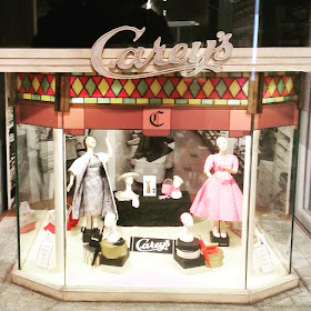 Model of a draper's front window with mannequins displaying frocks and hats. Several signs say 'Carey's'.