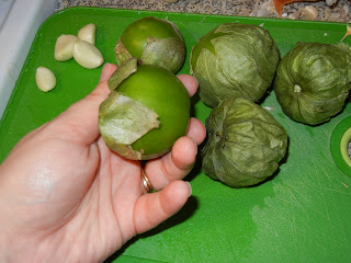 holding a peeled tomatillo and the rest of the tomatillos on unpeeled on a green cutting board with 4 cloves of peeled garlic to the top left of the green cutting board 
