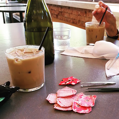Table in a cafe with two glasses of iced coffee and a bottle of water on it. Next to the drinks is a pile of miniature cushions, worng-side out, two pairs of tweezers, a length of white fabric and a quik-unpikc.