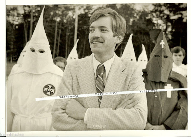 David Duke attending a Louisiana Ku Klux Klan rally in 1979 (Photograph by Michael P. Smith). A mustachioed Duke is in the foreground wearing a light colored tweed suit and striped tie. His arms folded, he is grinning, and looking off into the distance. Behind him are several hooded and draped members of the Ku Klux Klan.