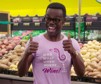 Chidi, standing in front of a supermarket display of multiple types of potatoes, is wearing a light purple shirt that says "WHO/WHAT/WHEN/WHERE... Wine!" Next to the writing, there's a line drawing of a wineglass with a swirl of wine splashing out the top.