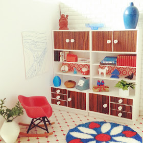 One-twelfth scale miniature retro lounge with white walls, woodgrain and white display units, red Eames rocking chair and a round retro rug in colours of red, teal and white on the floor.