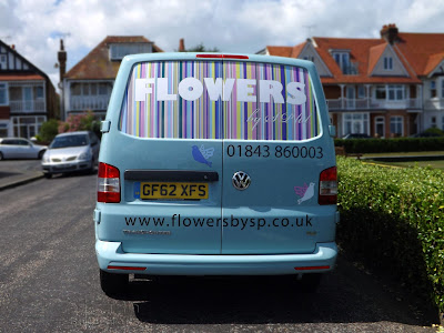 Back of an eggshell coloured Volkswagen van, with a large window with stripes in pastel shades of pink, blue, green and yellow, Flowers in a bold white font, with a script text 'by SP ltd' underneath. Two doves on the body of the car in blue, white and pink. Vinyl lettering for the phone number and website address.