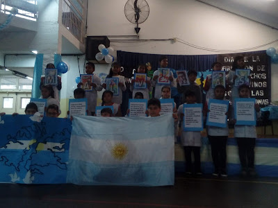 se ven de pie a lños alumnos de quinto grado sosteniendo afiches con fragmentos de la poesía. También sostienen una Bandea Argentina y otra de Malvinas