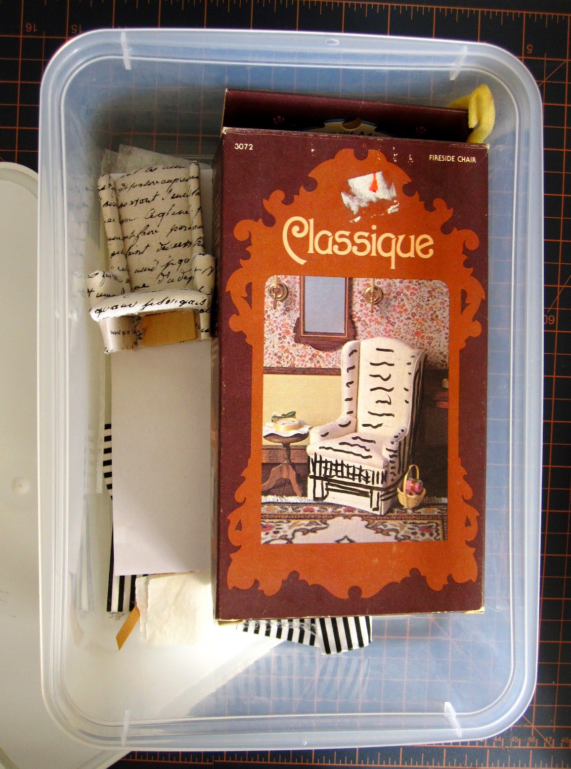 Plastic storage box containing a dolls' house miniature wing chair kit package, a half-finished chair and various pieces of fabric.