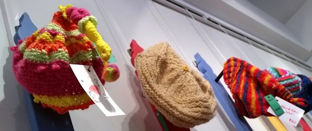 Three brightly coloured hats displayed on the southern wall of the exhibition. Left: long 'sleeping cap' style of beanie in candy colours. Centre a beret style knitted hat in a neutral beige; Right: a pointed knitted beanie in purple, yellow and hot pink stripes and ribbed band.