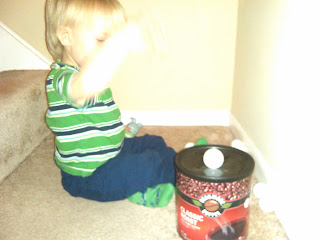 toddler boy sitting on bottom of stairs playing with plastic golf ball and coffee can homemade shape sorter