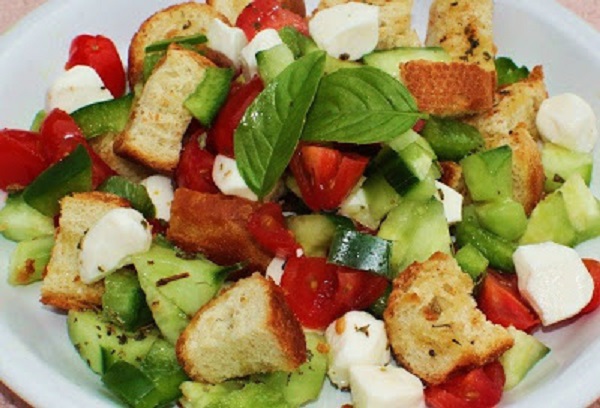 this is a salad in a white plate with  fresh basil, cucumbers, cubed mozzarella, toasted bread cubes and tomatoes all fresh